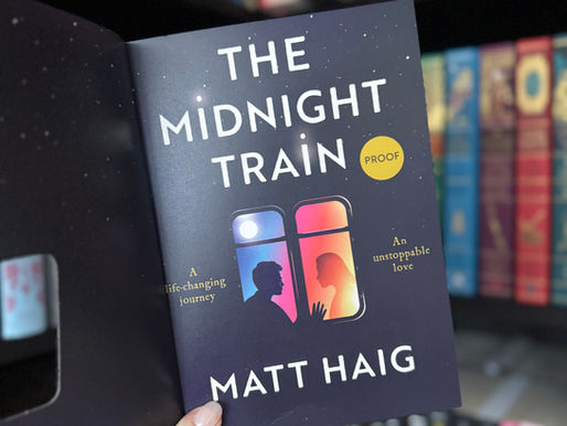 Hand holds "The Midnight Train" proof by Matt Haig, featuring silhouettes and vibrant colors. Bookshelf with colorful books in the background.