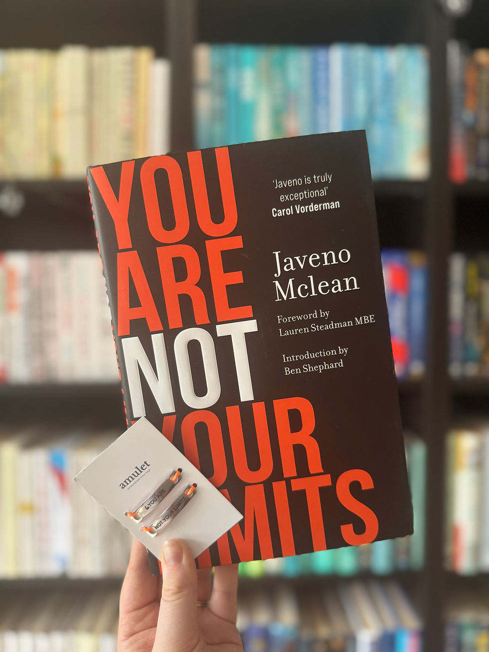 Hand holds a black book with bold red text "YOU ARE NOT YOUR LIMITS" by Javeno Mclean. Background: blurred colorful bookshelf.