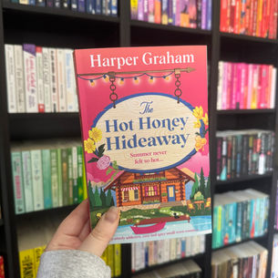 Hand holding The Hot Honey Hideaway by Harper Graham in front of colorful bookshelves.