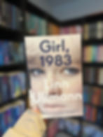 Person holding "Girl, 1983" by Linn Ullmann in a library. Blurred bookshelf background with colorful books. Focus on book cover.