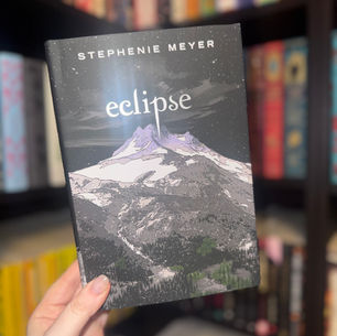 Hand holds "Eclipse" by Stephenie Meyer, showing a mountain scene cover. Background features a colorful, blurred bookshelf.