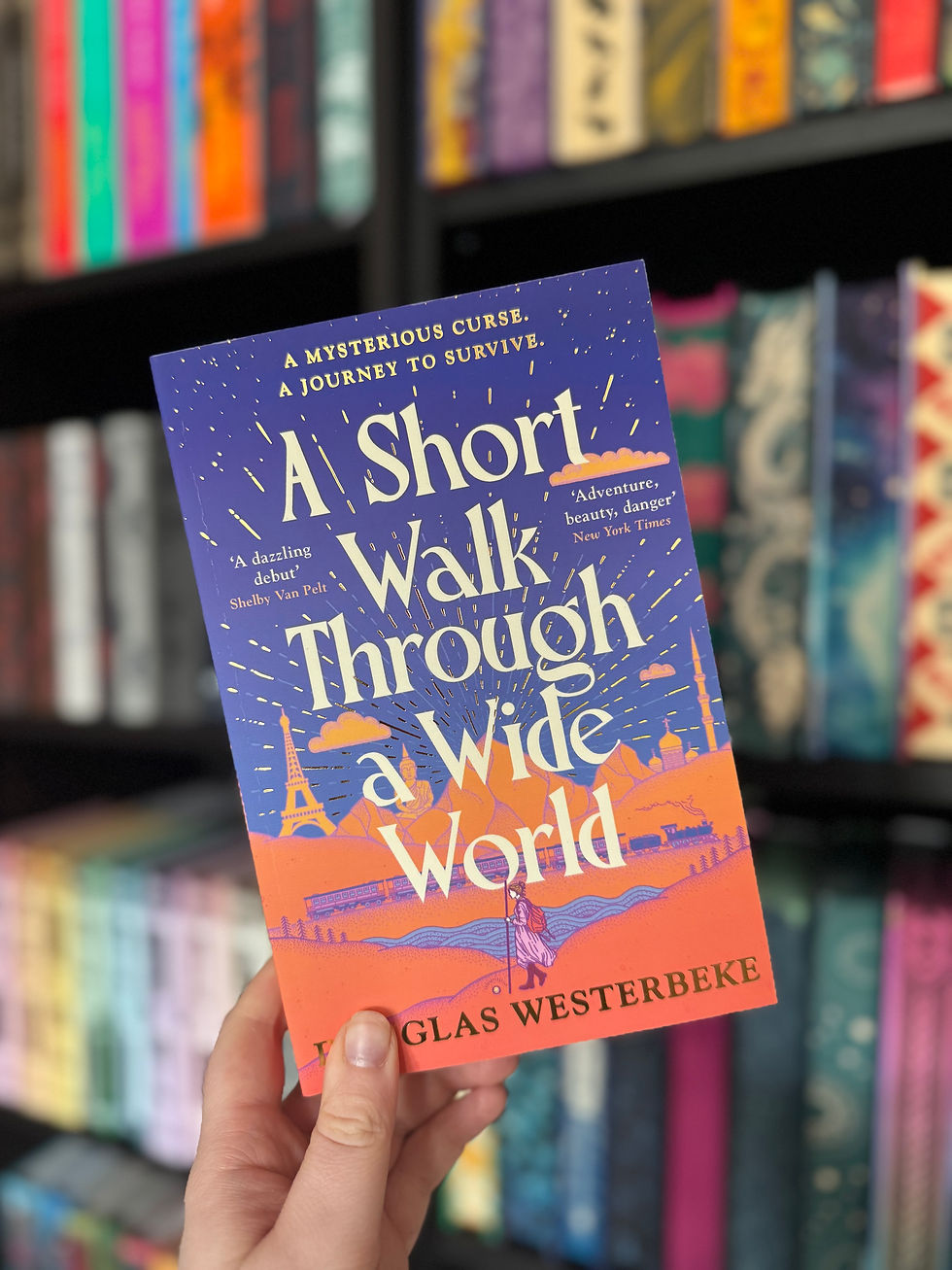 Hand holding a colorful book titled "A Short Walk Through a Wide World" by Douglas Westerbeke. Bookshelves with colorful spines in the background.