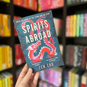 Hand holding a book titled "Spirits Abroad" by Zen Cho, featuring a red dragon on the cover. Background shows colorful, blurred bookshelves.