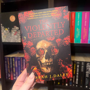 Hand holding the book "The Violently Departed" by Sarah J. Daley, featuring a skull with red flowers, in front of colorful bookshelves.