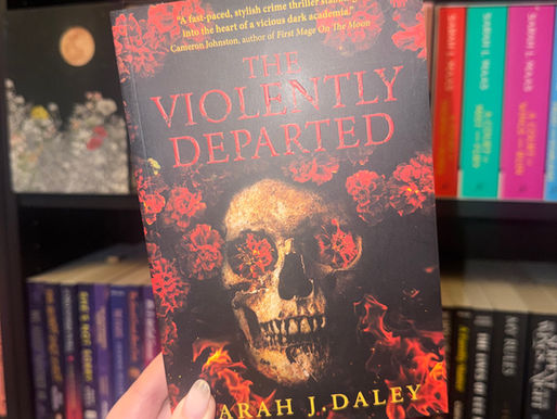 Hand holding the book "The Violently Departed" by Sarah J. Daley, featuring a skull with red flowers, in front of colorful bookshelves.