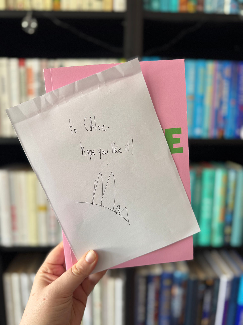 Hand holds a pink book with a note saying "To Chloe, hope you like it! Max." Blurry bookshelf in the colorful background.