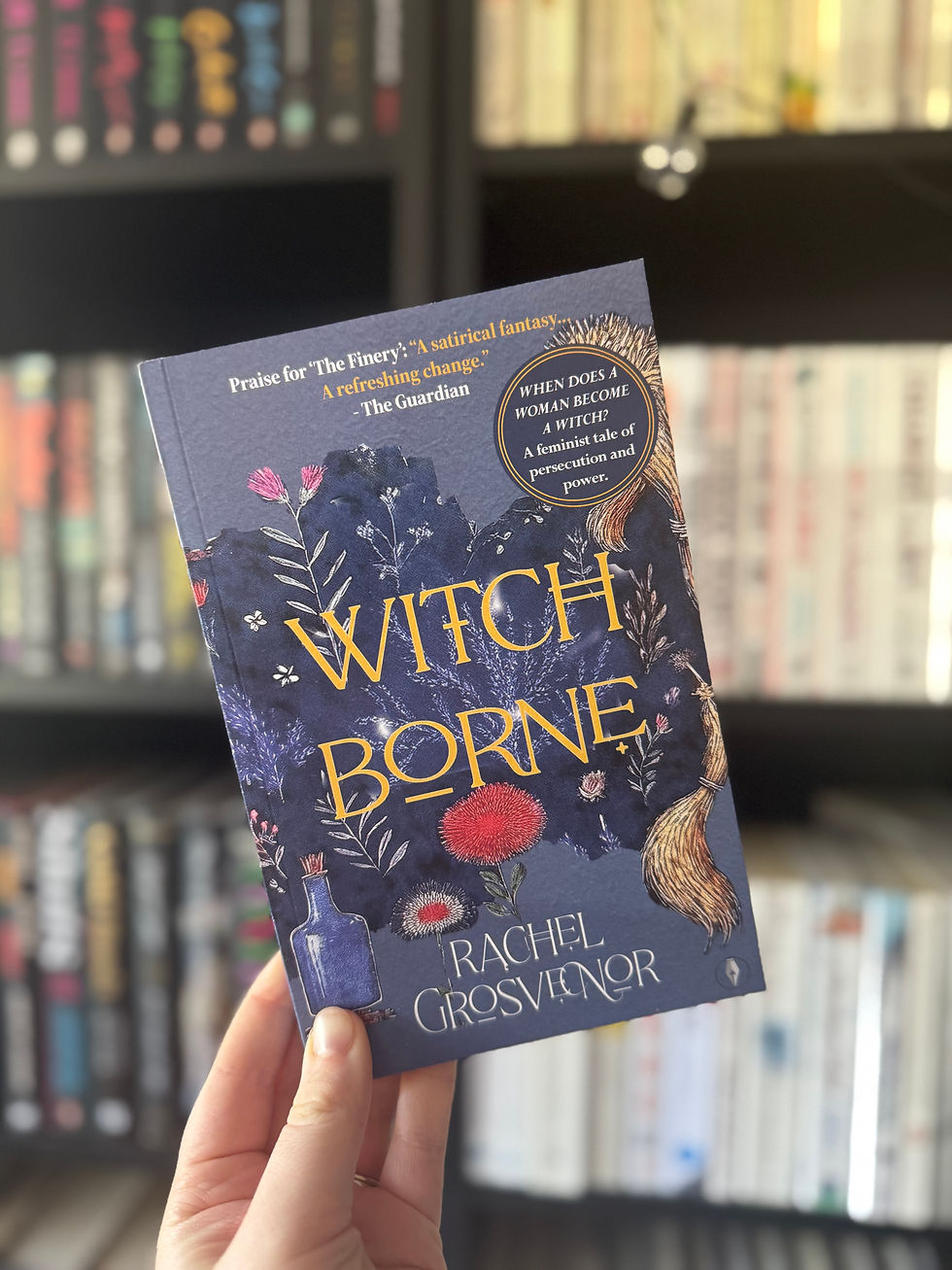 Hand holding the book "Witch Borne" by Rachel Grosvenor, with floral design and broom on cover. Shelves of colorful books in the blurred background.