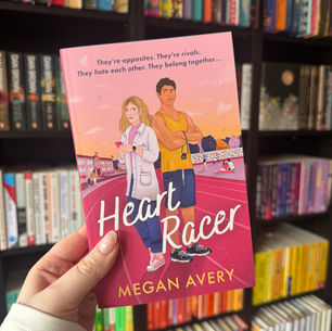 Person holding a pink book titled "Heart Racer" by Megan Avery, featuring illustrated characters. Bookshelves with colorful books in background.