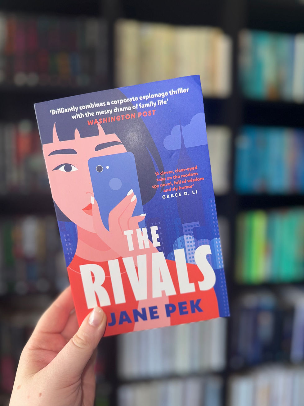 Hand holds "The Rivals" by Jane Pek, featuring a stylized woman with a phone on the cover. Bookshelf blurred in the background.
