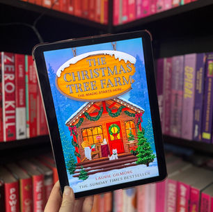 Hand holding a tablet displaying "The Christmas Tree Farm" book cover with a festive cabin. Colorful bookshelves in the background.