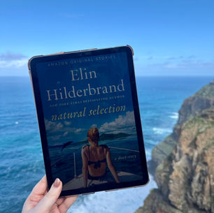 Tablet showing "Elin Hilderbrand, Natural Selection" held by a hand. Background: ocean and cliffs under blue sky. Calm and scenic.