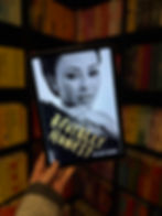 A hand holds a tablet displaying "Brutally Honest" by Melanie Brown. The background shows colorful, blurred bookshelves.