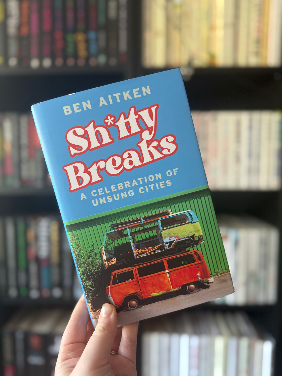 Hand holding a book titled "Sh*tty Breaks" by Ben Aitken, with a blue cover featuring a stacked bus. Bookshelf background, colorful spines.