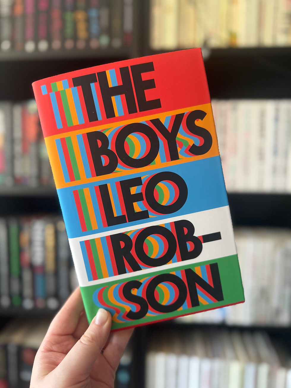 Hand holding a vibrant book titled "The Boys" by Leo Robson, with bold colors and patterns. Blurred bookshelves in the background.