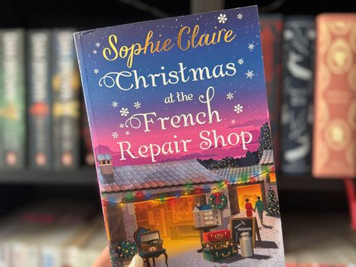 Hand holds a book titled "Christmas at the French Repair Shop" by Sophie Claire. Cozy festive illustration. Bookshelves in the blurry background.