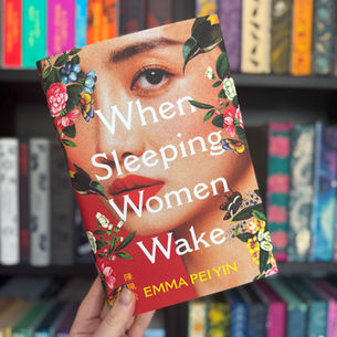 Hand holding a colorful book titled "When Sleeping Women Wake" by Emma Pei Yin. Vibrant floral cover. Bookshelf background.