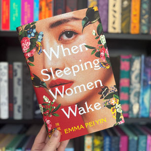 Hand holding a colorful book titled "When Sleeping Women Wake" by Emma Pei Yin. Vibrant floral cover. Bookshelf background.