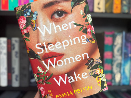 Hand holding a colorful book titled "When Sleeping Women Wake" by Emma Pei Yin. Vibrant floral cover. Bookshelf background.