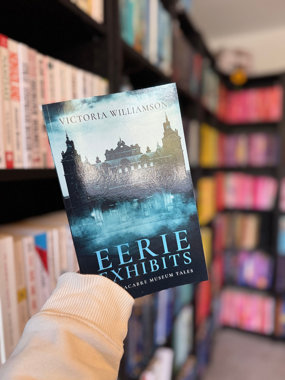 Hand holding a book titled "Eerie Exhibits" by Victoria Williamson in a colorful library setting. Bookshelves in the blurred background.