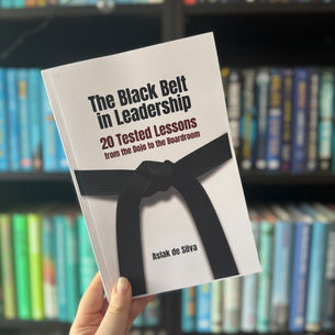 Hand holding a book titled "The Black Belt in Leadership" by Aslak de Silva, set against a colorful bookshelf background.