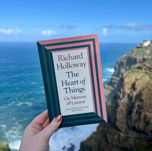 Hand holding a book titled "The Heart of Things" by Richard Holloway against a cliffside and ocean backdrop under a clear blue sky.