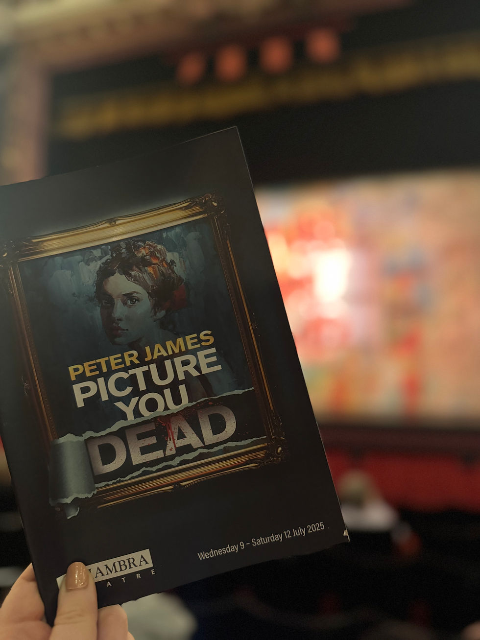 A hand holds a theater program titled "Peter James: Picture You Dead" in a dimly lit theater, with blurred red seats in the background.