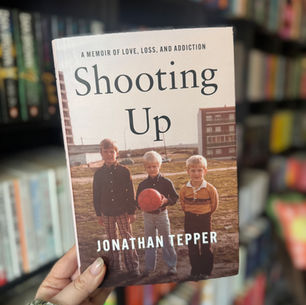 A hand holds the book "Shooting Up" by Jonathan Tepper, featuring a cover photo of three boys with a ball. Bookshelves are in the background.