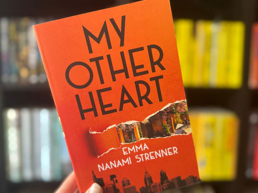 Hand holds an orange book titled "My Other Heart" by Emma Nanami Strenner, with a blurred bookshelf background filled with colorful books.