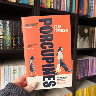 Hand holding "Porcupines" book by Fran Fabriczki in a colorful bookstore. The cover is orange with an illustration of a woman and girl.
