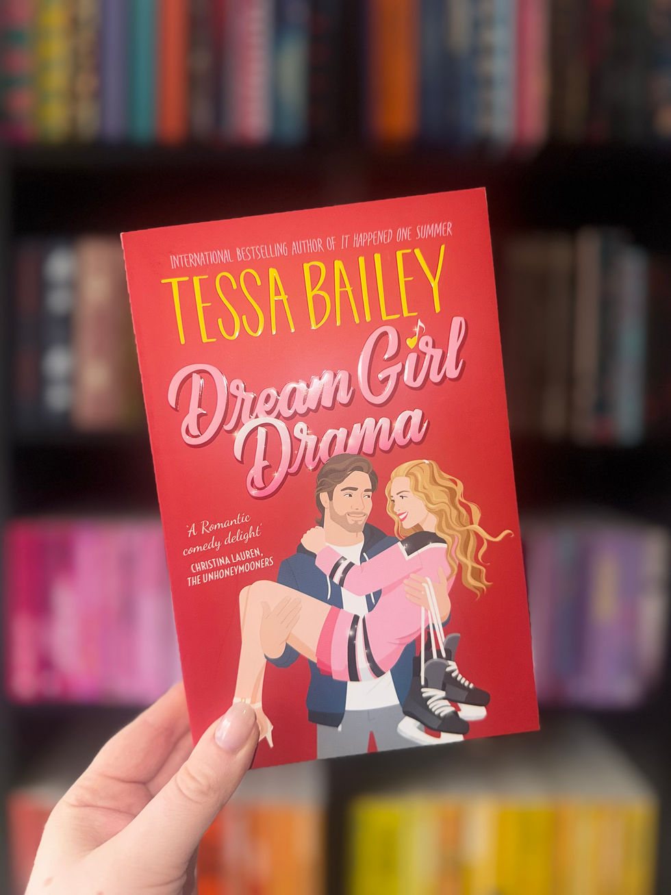 Hand holds a book titled "Dream Girl Drama" by Tessa Bailey. The colorful cover shows a man holding a woman. Blurred bookshelves in the background.
