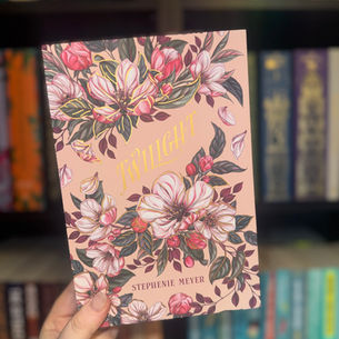 A hand holds a "Twilight" book by Stephenie Meyer with a floral cover. Blurred colorful books are in the background.