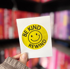 A hand holds a retro-style yellow sticker with a smiley face and the text "BE KIND REWIND" in black letters. The background features a colourful bookshelf arranged by spine colour, with red, pink, and purple tones visible.