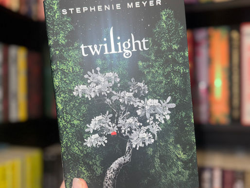 A hand holds the book "Twilight" by Stephenie Meyer. The dark cover features a white tree with leaves and a red apple. Bookshelf background.