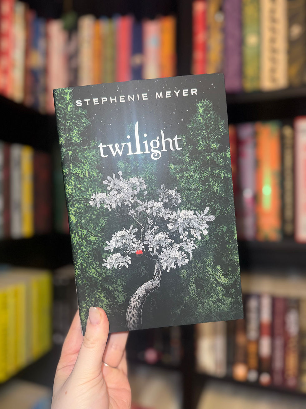 A hand holds the book "Twilight" by Stephenie Meyer. The dark cover features a white tree with leaves and a red apple. Bookshelf background.