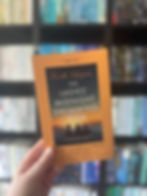Hand holds "The Ladies’ Midnight Swimming Club" book against a blurred bookshelf background. Aqua, orange, and black cover with ocean scene.