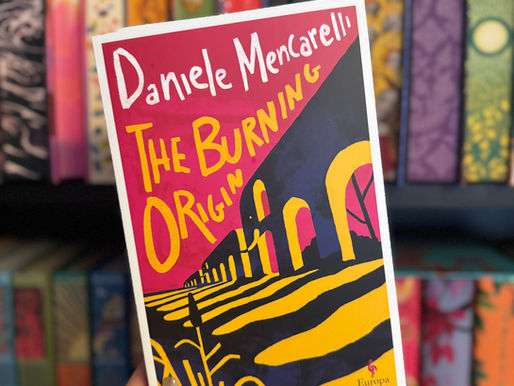 Hand holding "The Burning Origin" book by Daniele Mencarelli, with colorful cover. Bookshelf with vibrant spines in the background.