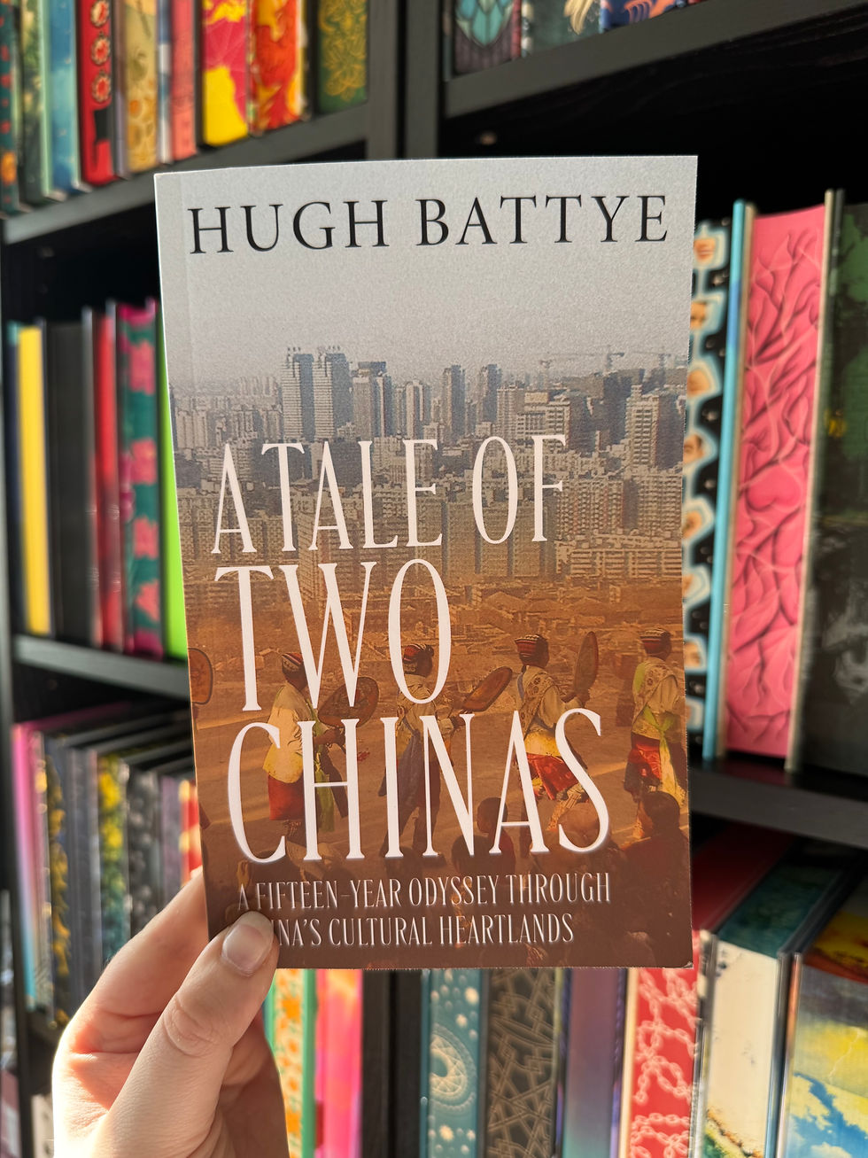 Hand holding Hugh Battye’s A Tale of Two Chinas in front of a colorful bookshelf.