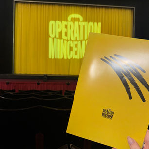 Bright yellow theater program for "Operation Mincemeat" held in front of a matching curtain with the same title; empty seats visible.