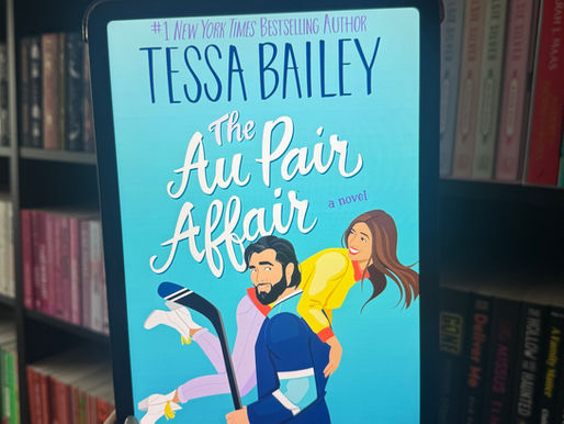 E-reader displaying "The Au Pair Affair" by Tessa Bailey, held by a hand. Bright blue cover with illustrated couple. Background: bookshelves.