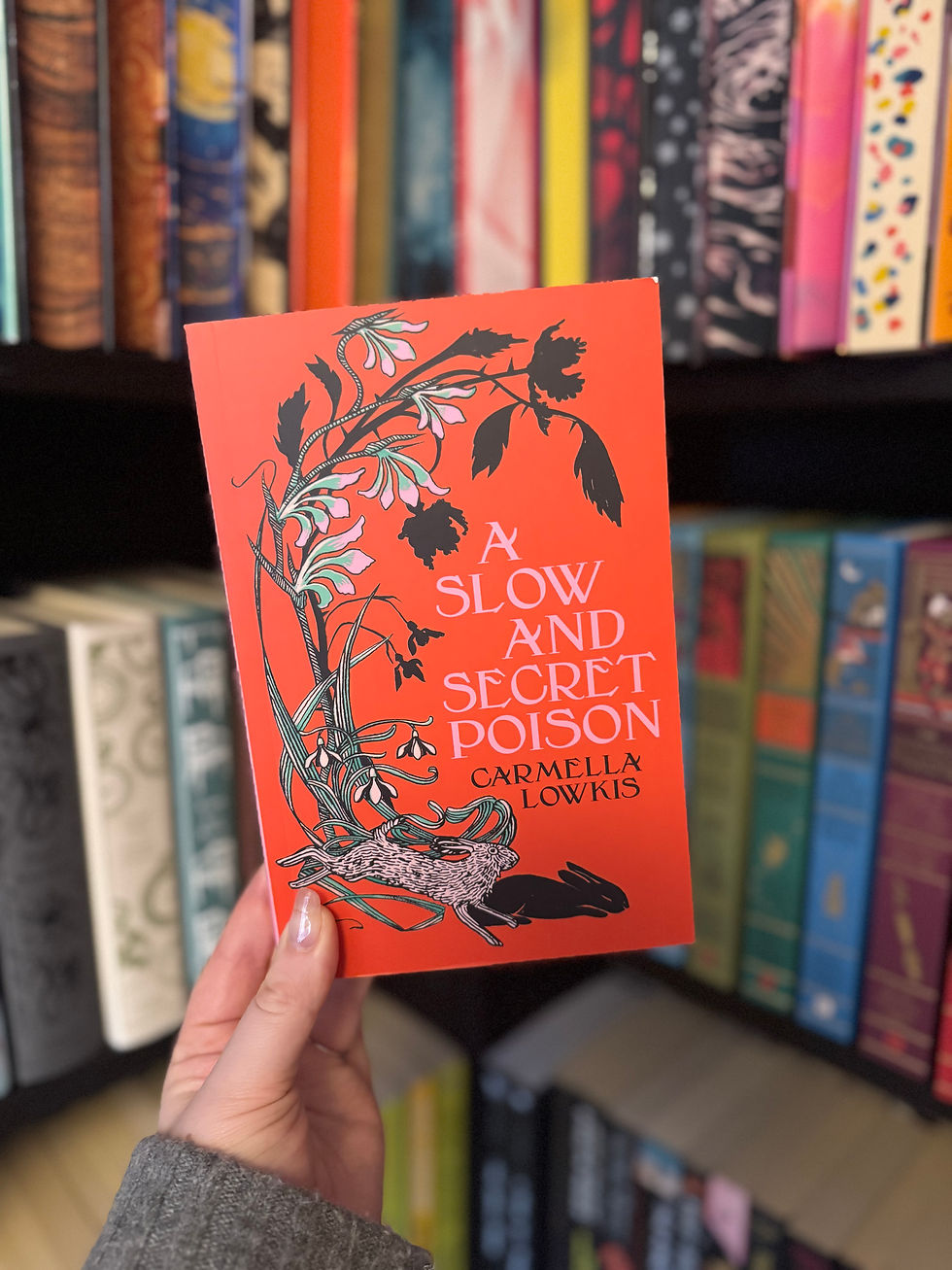 Hand holding an orange book titled "A Slow and Secret Poison" by Carmella Lowkis, with botanical art, in front of a colorful bookshelf.