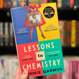 Hand holding the book "Lessons in Chemistry" by Bonnie Garmus against a bookshelf. Cover features a vintage TV illustration and vibrant colors.
