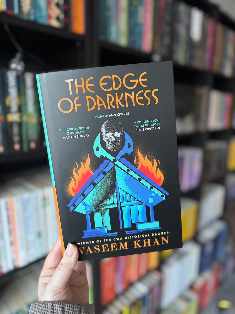 Hand holding "The Edge of Darkness" book with fiery house cover, by Vaseem Khan. Background shows colorful bookshelves, creating a vibrant mood.