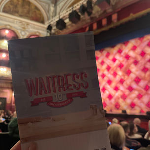 Hand holding a "Waitress" 10th anniversary program in an ornate theater. Red-patterned stage in the background. Audience seated.