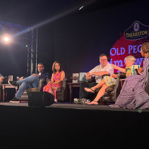 Panel of six people on stage discussing books, with bright spotlight and "Theakston" logo in the background. Participants are relaxed and engaged.