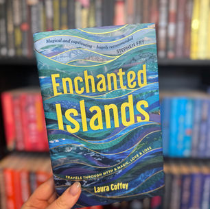 A hand holds a book titled "Enchanted Islands" by Laura Coffey. The cover is blue with wave patterns. Bookshelves are blurred in the background.