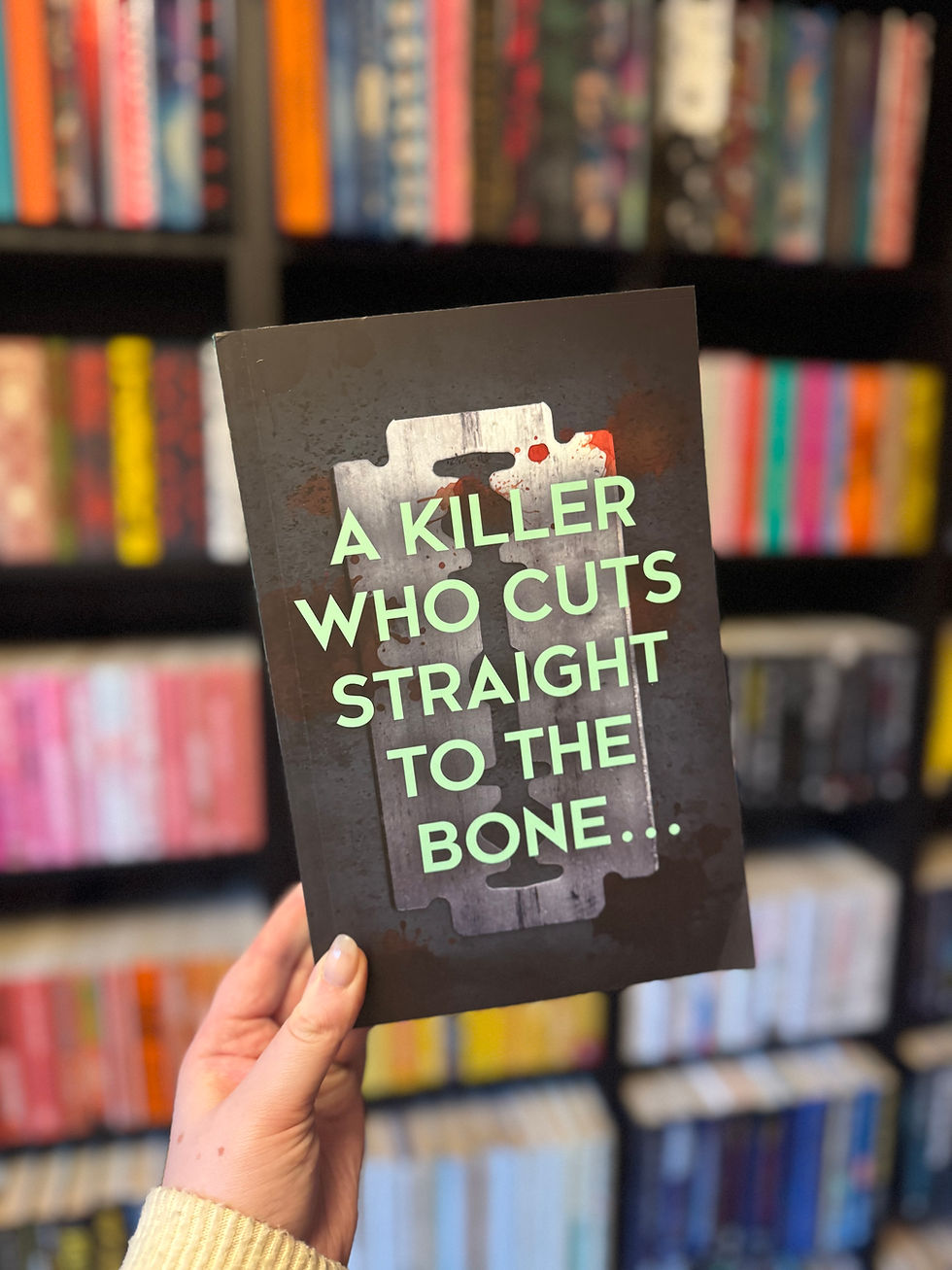 Hand holding a book with the text "A KILLER WHO CUTS STRAIGHT TO THE BONE..." against bookshelves in the background.