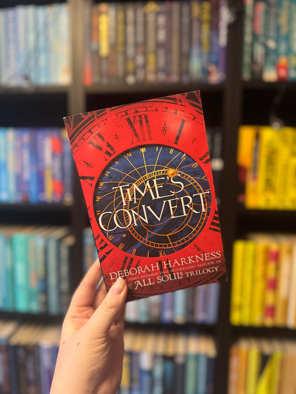 Hand holding "Time's Convert" book with red clock cover, set against a colorful bookshelf background, creating a cozy library vibe.