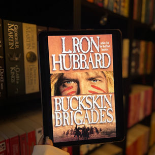 A hand holds a tablet displaying the book cover "Buckskin Brigades" by L. Ron Hubbard. Bookshelves full of colorful books are in the background.