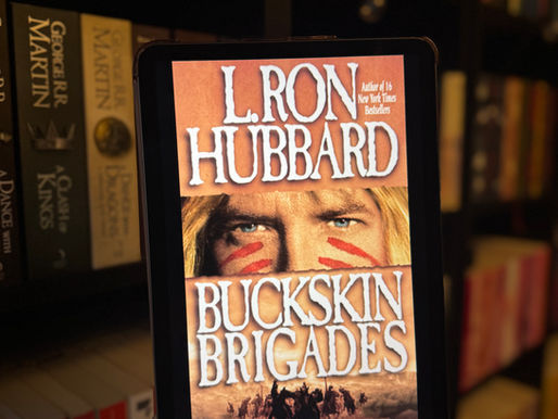 A hand holds a tablet displaying the book cover "Buckskin Brigades" by L. Ron Hubbard. Bookshelves full of colorful books are in the background.