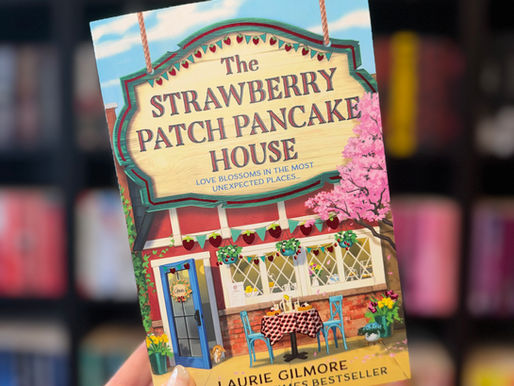 Hand holding "The Strawberry Patch Pancake House" book with colorful cover featuring a cozy café scene. Blurred bookshelves in background.